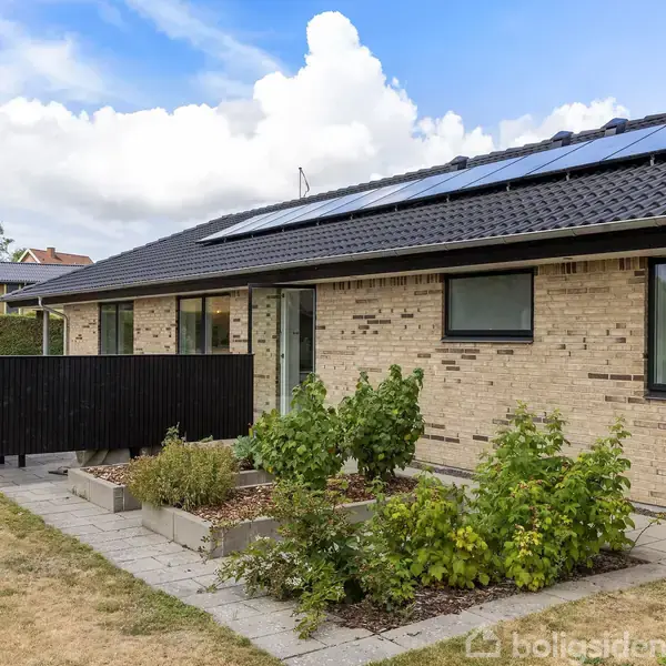 En étplans villa med solpaneler på taget. Bygningen har gul murstensfacade, omgivet af en have med fliser og buske. Himlen er blå med skyer i baggrunden.