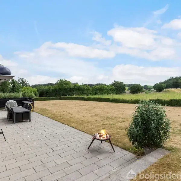 Terrasse med fliser mod græsplæne. Bålsted brænder svagt ved siden af en busk. I baggrunden ses marker og træer under blå himmel.