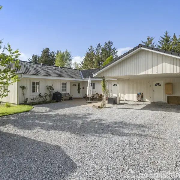 Et hvidt hus med sort tag står stille ved siden af en carport. De er omgivet af en grusbelagt gårdsplads og grønne træer.