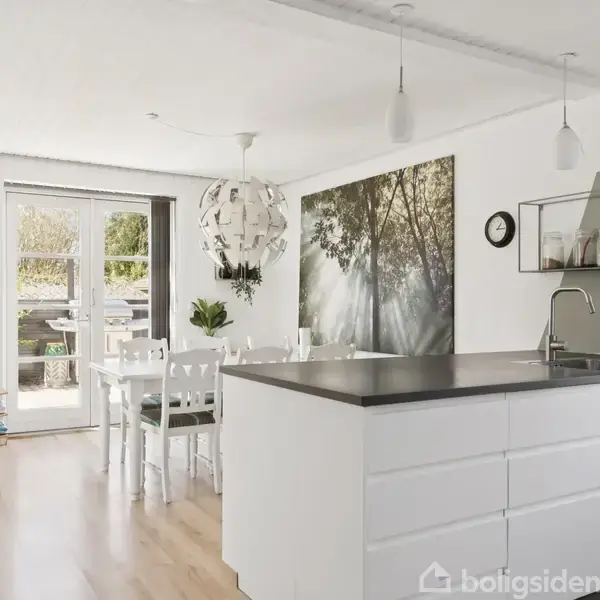 Køkkenø med hvid skabsoverflade i moderne køkken. Spisebord i baggrunden med stole foran en havedør. Vægdekoration viser trækroner. Pendellamper hænger fra loftet. Udendørsområde udenfor.