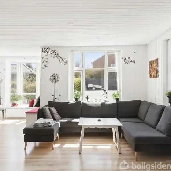 En stor, mørkegrå sofa står i en lys stue med trægulv og store vinduer. Væggen er dekoreret med blomsterdekaler, og der står en hvid rektangulær sofabord foran sofaen.