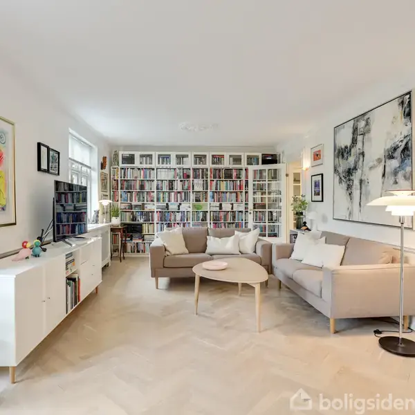 Stue med sofa og sofabord står foran en stor bogreol fyldt med bøger. Vægkunst og lamper skaber en hyggelig atmosfære. Planter og moderne indretning i rummeligt rum.