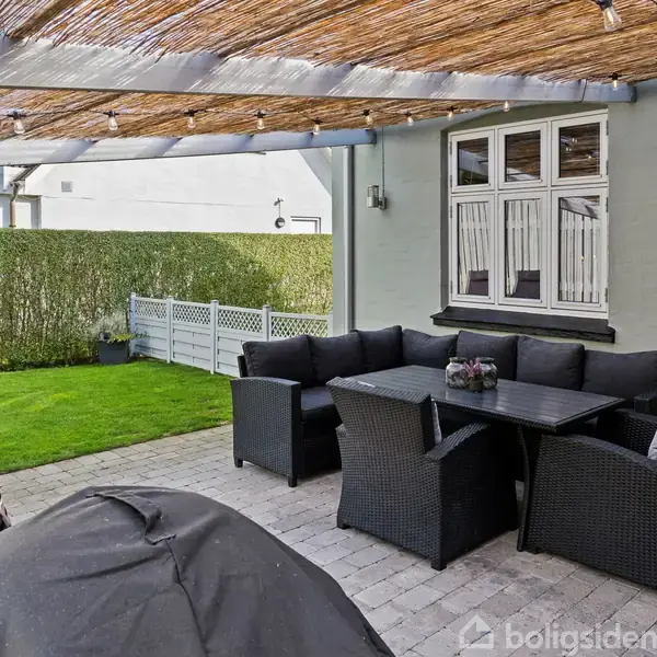 En udendørs terrasse med sorte kurvemøbler står under et halvtag med belysning. Omkring dem er en grøn græsplæne og en hæk. I baggrunden ses en hvid bygning.
