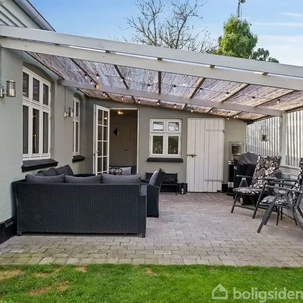 Terrasse med grå sofa- og stolearrangement under en overdækket pergola. Beliggende ved husets sidevæg med en grøn græsplæne foran. Møblerne vender mod et bord og en dør.