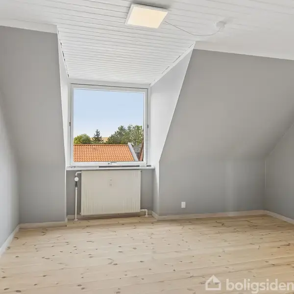 Et tomt værelse med lysegrå vægge og trægulv, har et vindue med udsigt til et tag og træer. Et hvidt loft med en lampe hænger over rummet.