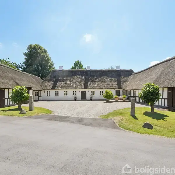 Bindingsværkshus med stråtag danner gårdsplads. Huset har hvid facade med sorte bjælker. Gården er omgivet af græsplæner med små træer. Himlen er blå og solrig.