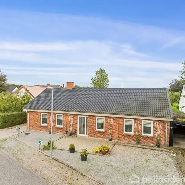 En rød murstensbygning med sort tag står stille ved en vej. Foran huset er der en parkeret bil og nogle blomsterkrukker. Omkring bygningen er der træer og andre huse.