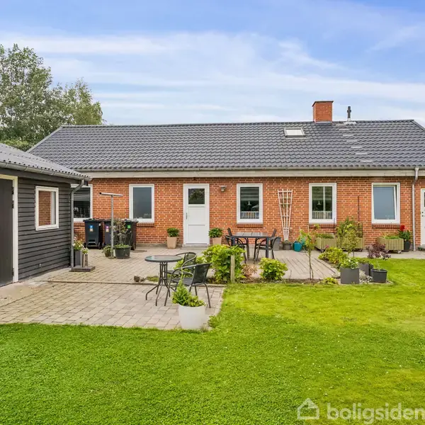 En murstensvilla står med en fliseterrasse i baggrunden og havemøbler. Græsplæne omgiver området, og en garage står til venstre. Himlen er let skyet og blå.