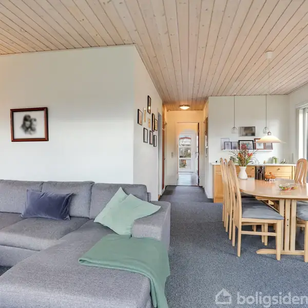 Gråt hjørnesofa med puder står i stue. Spisebord med træstole ses ved siden af. Rummet er dekoreret med hængelamper og billeder på væggene. Tæppet er mørkegråt.