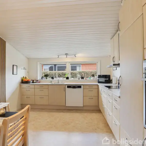 Et lyst køkken med hvide skabe og skuffer, en indbygget ovn og opvaskemaskine i midten. Et vindue bagved åbner mod udsigt, og spisebord med stole ses til venstre.