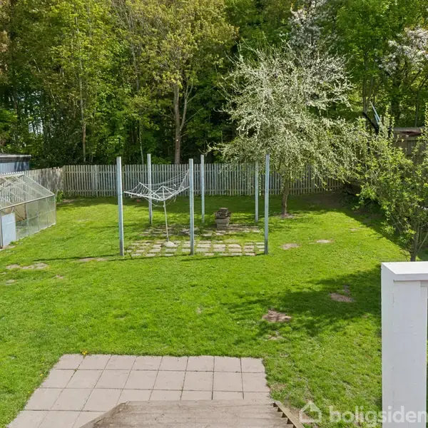 Græsplæne med tørrestativ, omgivet af et hegn og træer. Et drivhus står i venstre side, mens et flisebelagt område leder fra en terrasse.