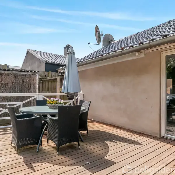 Terrassen har et rundt bord med fire stole og en parasol. Bordet står på et trægulv ved siden af en lys væg med en skydedør. Atmosfæren er udendørs og afslappet.