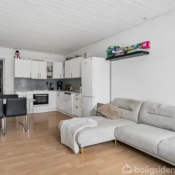 Grå sofa står i en åben stue-køkken-alrum, hvor et hvidt køkken med sort-hvid mønstrede fliser ligger i baggrunden. To sorte spisebordsstole står ved en lille bordplade.