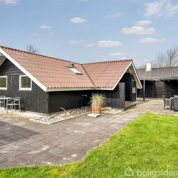 Et sort træhus med rødt tegltag står stille omgivet af fliser og en grøn græsplæne. En grill og havemøbler ses på terrassen under en lyseblå himmel.