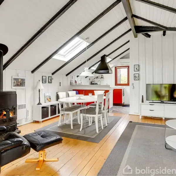 En hyggelig stue med en brændeovn i hjørnet, hvide vægge og loft med sorte bjælker. Et spisebord med hvide stole står på et tæppe, omgivet af forskellige møbler og en TV.