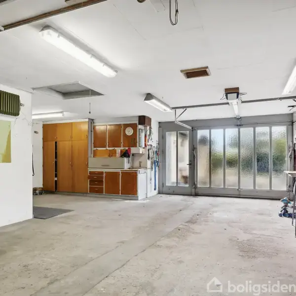 Garage med tomt beton gulv, værktøjsreoler og arbejdsborde langs væggene. Bagtil er der glasporte med udsigt til en have. Overlysbelysning hænger fra loftet.