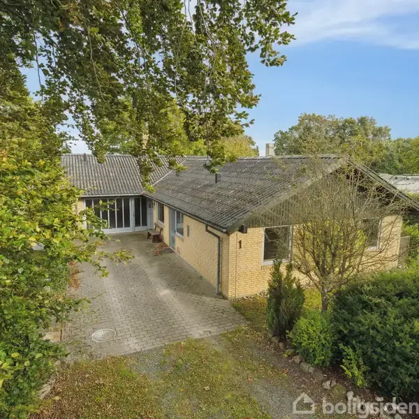 Et gult murstenshus står stille omgivet af frodige træer og en grusbelagt indkørsel. Baggrunden viser en klar blå himmel med spredte skyer.