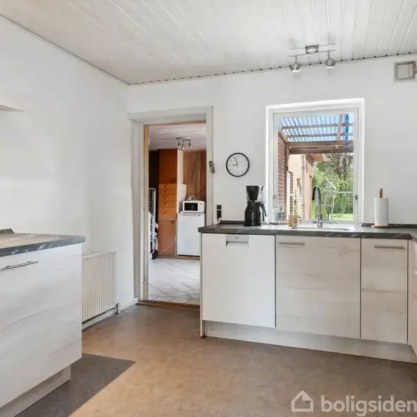 Et lyst køkken med hvide skabe og sorte bordplader. En kaffemaskine står på bordet ved et vindue, der fører ud til en have. En dør åbner til en anden rum.