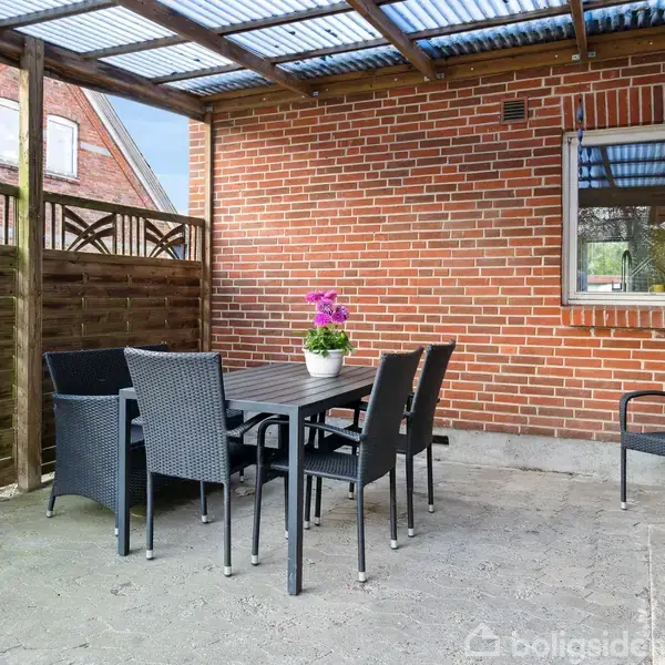 En udendørs terrasse med et bord og seks stole står på betonbelægning. Overdækket af et klart tag, omkranset af en murstensvæg og hegn. Blomster hænger og står i potter.