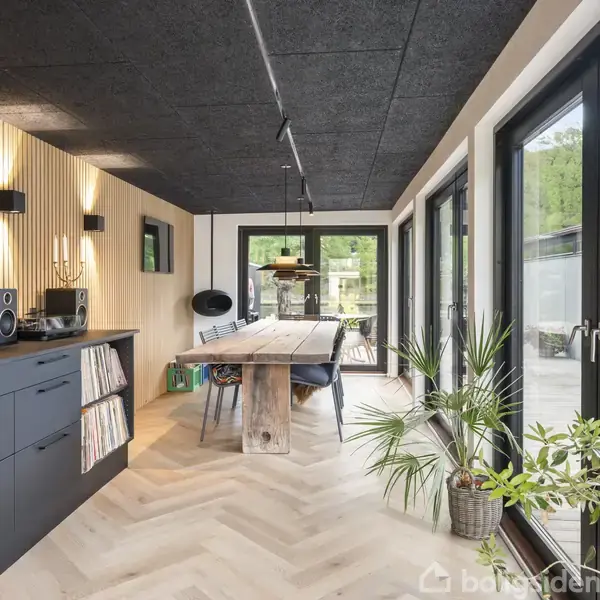 Stue-køkken-område med sort skabsmodul og kaffemaskine til venstre, et langt spisebord centralt. Store vinduer til højre giver udsigt til en have. Golv i fiskebensmønster.