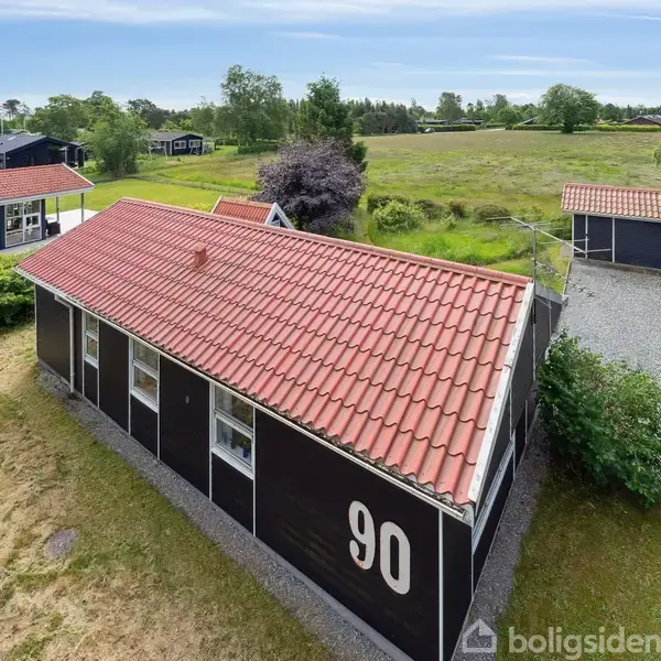 Et sortmalet hus med rødt tegltag har tallet "90" på siden. Det står i en grønt område med træer og buske, og en mindre bygning ses i nærheden på grusvej.