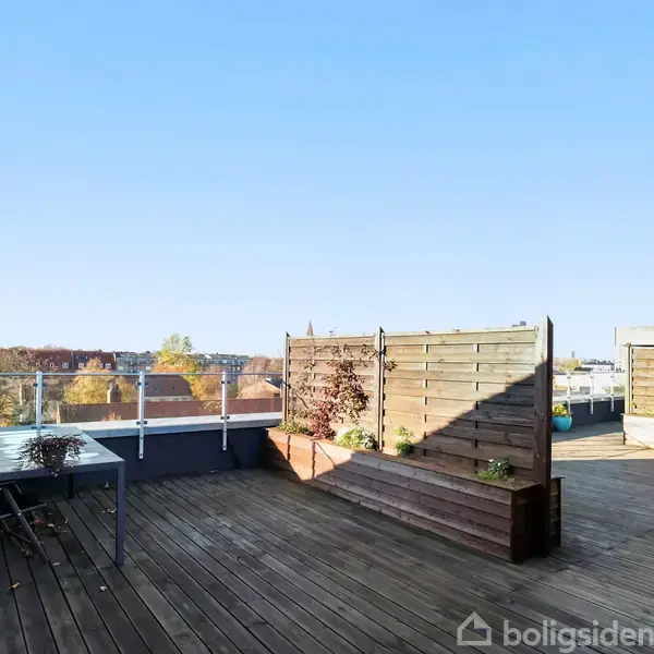 En træterrasse med bord og stole står stille under en klar blå himmel, omgivet af glasrækværk og plantekasser. I baggrunden ses byens tage og efterårsfarvede træer.