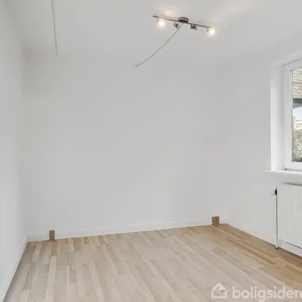 Et tomt rum med lyse trægulve og hvide vægge har en lampe i loftet og et stort vindue til højre der giver udsigt til en bygning udenfor.