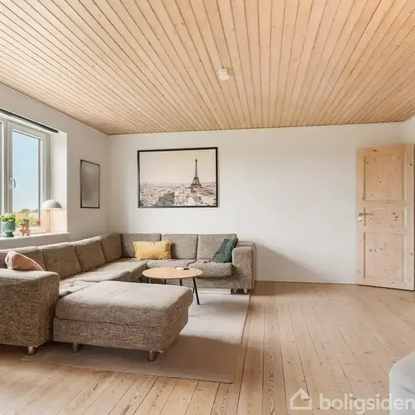 En stor, beige hjørnesofa står i en lys stue med trægulv og træloft. Et stort vindue til venstre giver udsigt, og en sort-hvid plakat hænger på væggen.