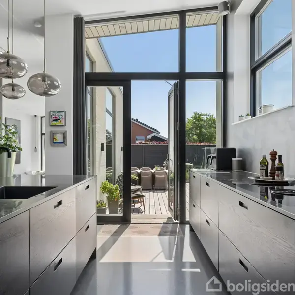 Et moderne køkken med sorte overflader fører ud til en terrasse med borde og stole. Store vinduer lukker masser af lys ind, og planterne giver et grønt touch.
