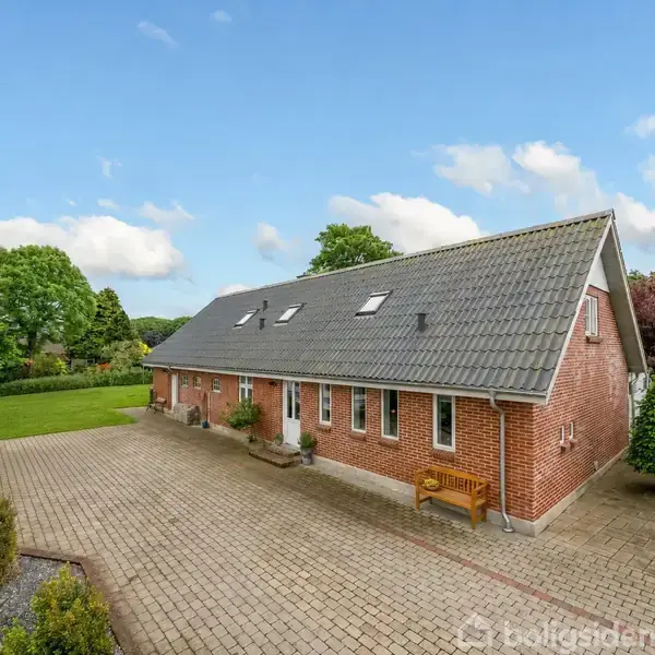 En rød murstensbygning med gråt tag står i en velholdt have. Der er en flisebelagt gårdsplads foran huset. Omkringliggende træer og buske skaber en frodig atmosfære.