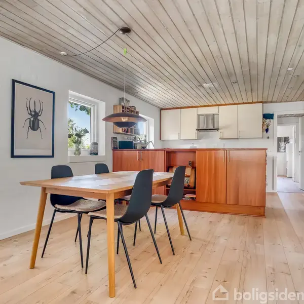 Et spisebord med sorte stole står i et lyst køkken-alrum. Over bordet hænger en lampe. Væggene er dekoreret med billeder. Køkkenskabe i træ ses i baggrunden.