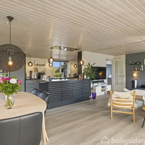 Et moderne køkken-alrum med åbent loft og trægulve. Bordet er dekoreret med blomster. Køkkenet har sorte skabe. Plakater og planteskrukker pynter rummet, som har et sofahjørne i baggrunden.
