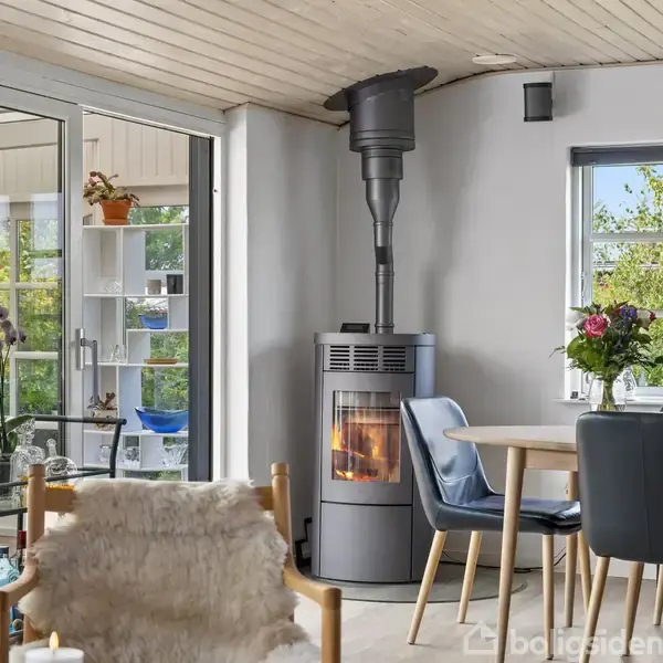 Brændeovn opvarmer stuehjørne med spisebord og stole i forgrunden, mens store vinduer og glasdør viser en grøn have i baggrunden; dekoreret med planter og lamper.
