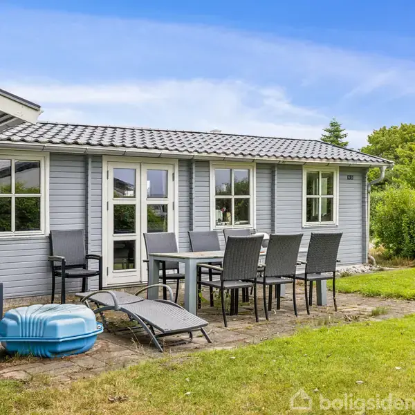 Et gråt træhus med hvide vinduer står i en frodig have. En terrasse foran huset har et sort bord og seks stole. En turkis plastikboks er placeret ved siden af en liggestol.