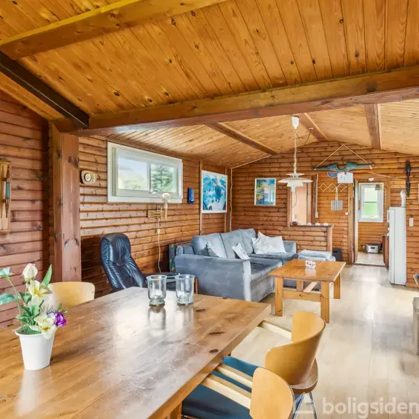 Et træbeklædt rum med spisebord og stole, sofaområde med blå sofa, og dekorationer på væggene. Store vinduer giver lys, og en dør fører til et andet rum.