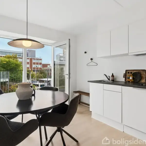 Et moderne, lyst køkken med en sort rund spisebord og fire stole, beliggende nær en stor glasdør, som fører ud til en balkon med udsigt til en bygning.