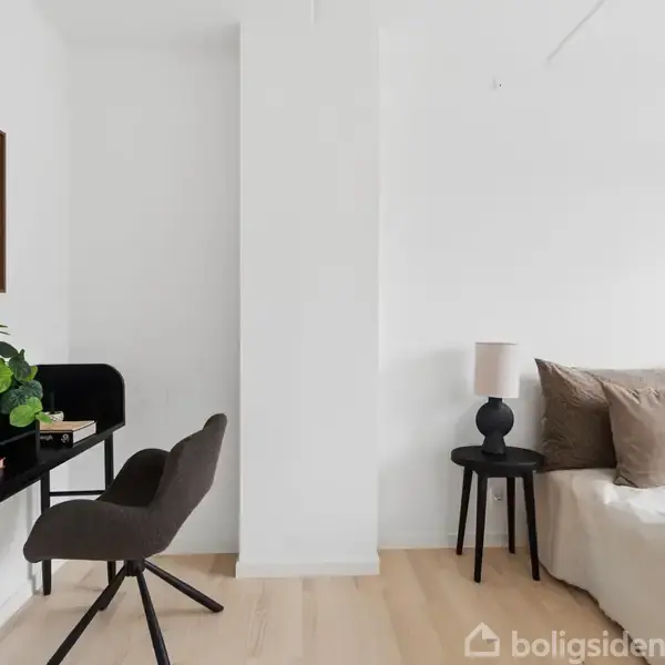 Et skrivebord med en plante og en stol står tæt ved en hvid væg. Til højre er en seng med puder og et lille bord med en lampe i en lys, minimalistisk indretning.