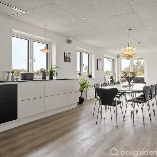 Et moderne køkken-alrum med sorte og hvide skabe. Rundt spisebord med sorte stole i midten. Store vinduer lader lys strømme ind, og planter står langs vinduerne.