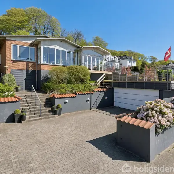 Et moderne hus med store vinduer og terrasse ligger på en skråning omgivet af træer og buske i blomst. En dansk flagstang står i indkørslen ved siden af en garage.