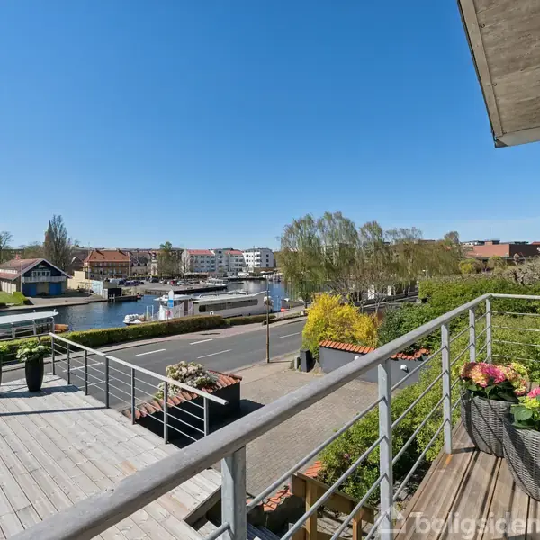 Balkon med blomster i kurve, vender ud mod en kanal med både. Solrig dag med klar blå himmel. Moderne bygninger og en kran ses i baggrunden.