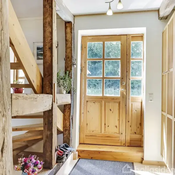 Trædør med glasruder står åben og slipper lys ind i en entré, omgivet af træbjælker og en trappe. Planter og blomsterdekorationer giver farve til rummet.
