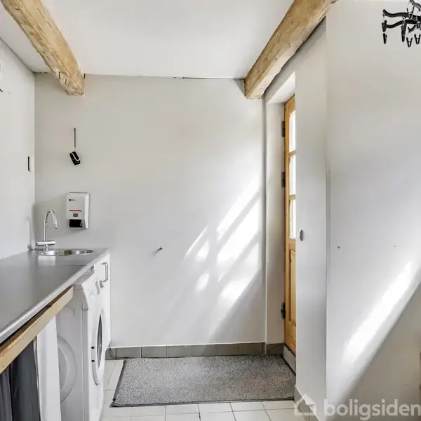 Vaskemaskine og tørretumbler står i et lyst bryggers med bjælker i loftet. Døren til højre slipper naturligt lys ind. Hylde med sko findes i hjørnet.