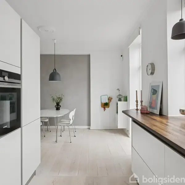 Et moderne køkken med hvide skabe og ovn; en træbordplade prydet med dekorationer. En spiseplads med hvidt bord og stole; store vinduer lukker naturligt lys ind.
