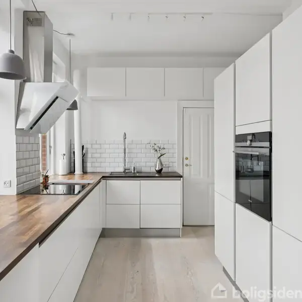 Et moderne køkken med hvide skabe og træbordplade. En indbygget ovn og emhætte er til stede. Store vinduer giver lys til rummet. Fliser bag vasken skaber et stilrent look.