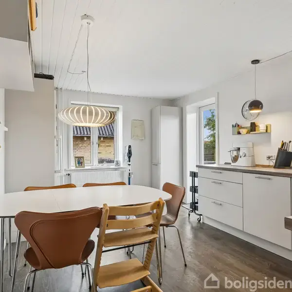 Spisebord med fire stole står i et lyst køkken. Moderne pendellamper hænger over bordet, omgivet af hvide skabe og store vinduer, der giver udsigt til en have.
