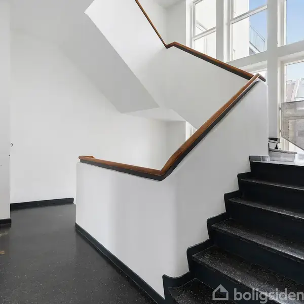 Trappe op mod store vinduer, hvor dagslyset strømmer ind. Hvide vægge og sorte trin skaber kontrast i rummets moderne design. En brun dørmåtte ligger ved siden af væggen.