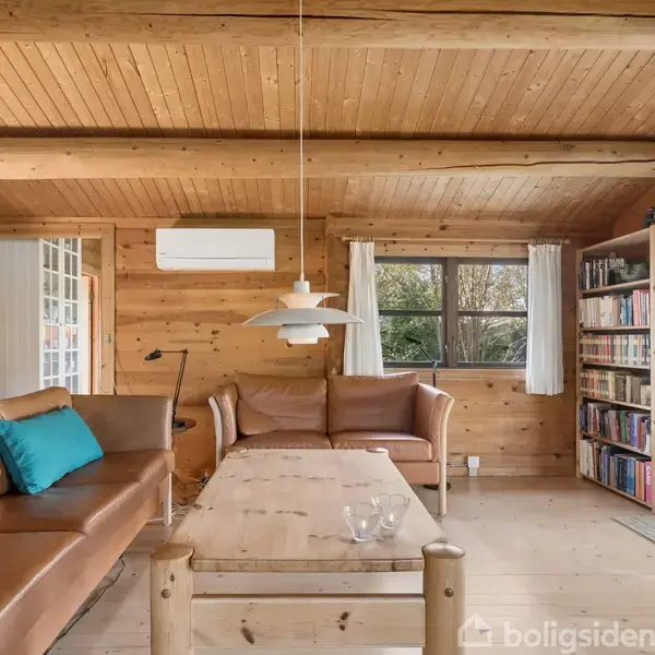 En stue med trævægge og loft har en brun lædersofa og et sofabord. En bogreol fyldt med bøger står ved siden af et vindue med hvide gardiner.
