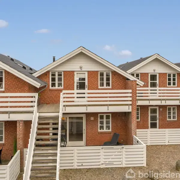 Røde murstens rækkehuse med hvidmalede altaner og trapper. Stenbelagt forhave med terrasse, omgivet af en blå himmel.