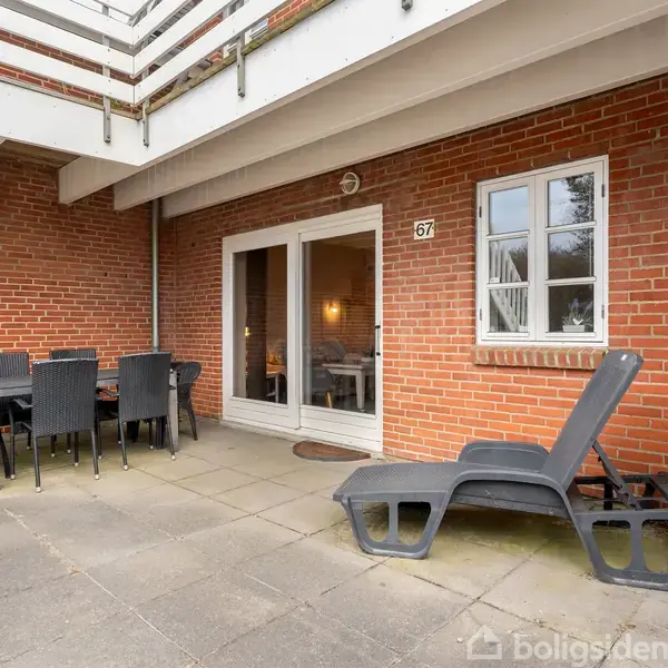 Terrasse med et bord og seks sorte stole står på fliser foran en rød murstensvæg. En liggestol er placeret ved siden af bordet. Nummer 67 ses ved døren.