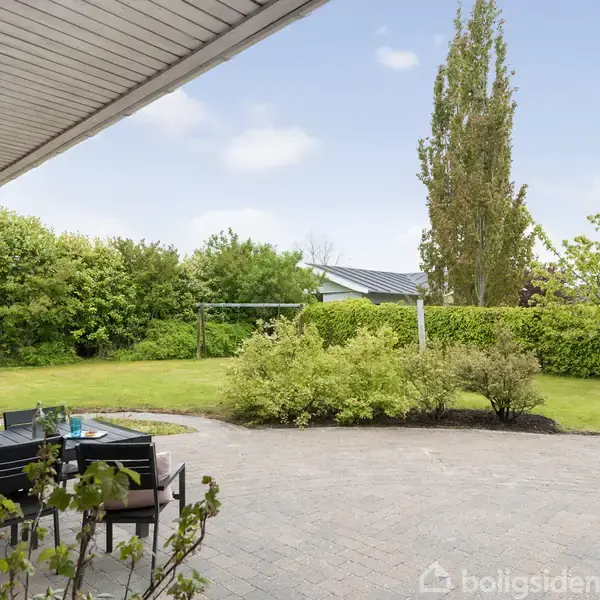 En fliseterrasse med havemøbler står ved siden af en velplejet græsplæne og buske i en baghave. I baggrunden ses en flagstang og forskellige træer under en blå himmel.